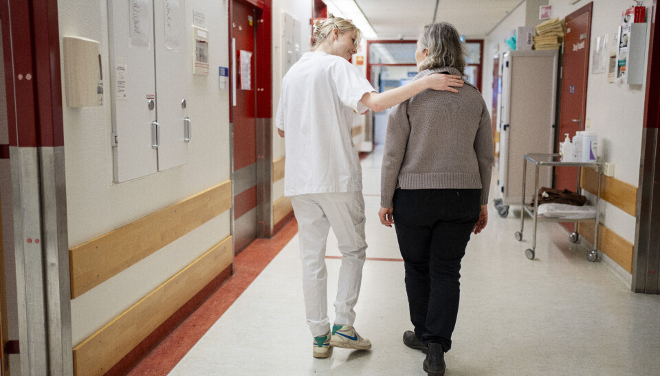 Meldingen anslår at vi kommer til å være 700.000 flere eldre i 2060 enn vi er i dag. Det øker behovet for arbeidskraft innen helse og omsorg. Kvinne i hvit helseuniform holder armen rundt eldre kvinne, begge går med ryggen til i en sykehuskorridor.