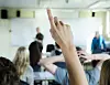 En hevet hånd i forgrunnen under undervisning i et klasserom, med elever og lærer i bakgrunnen.
